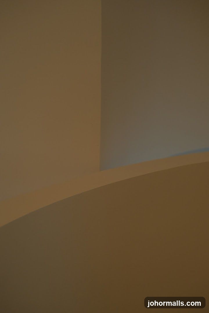 A minimalist abstract image featuring smooth, curved surfaces and sharp angles in muted tones of beige and brown. The lighting creates subtle shadows and highlights, emphasizing the architectural forms.