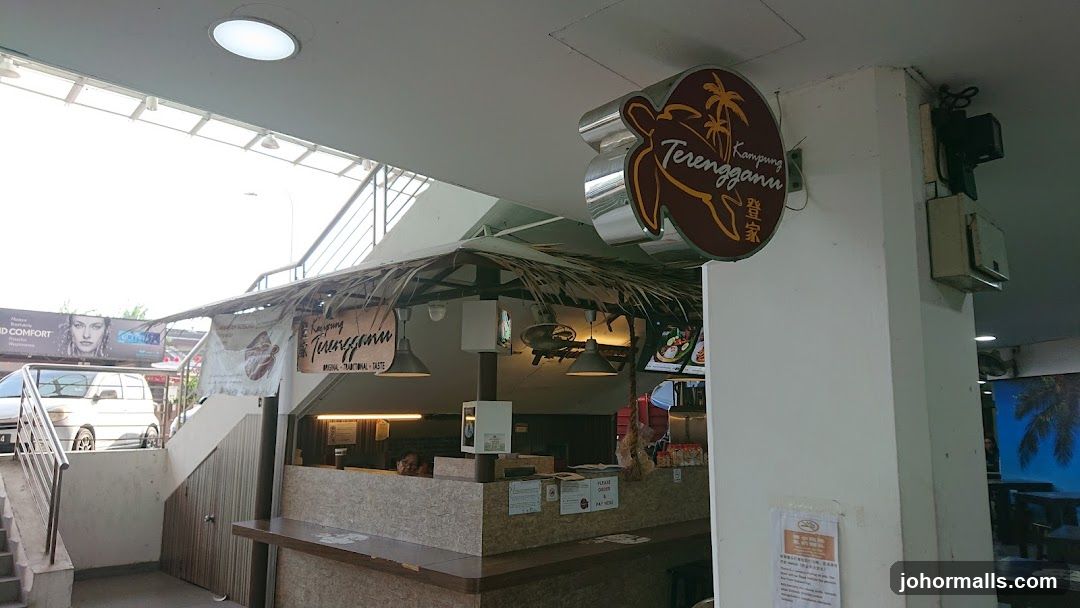 A view of the exterior of a food stall with a thatched roof. A prominent sign with a turtle logo and the text "Kampung Terengganu" is visible. Below the sign, another banner reads "Kampung Terengganu ORIGINAL. TRADITIONAL. TASTE." The stall has a counter with menus and what appear to be food displays. In the background, a large mural of a tropical beach scene is visible.