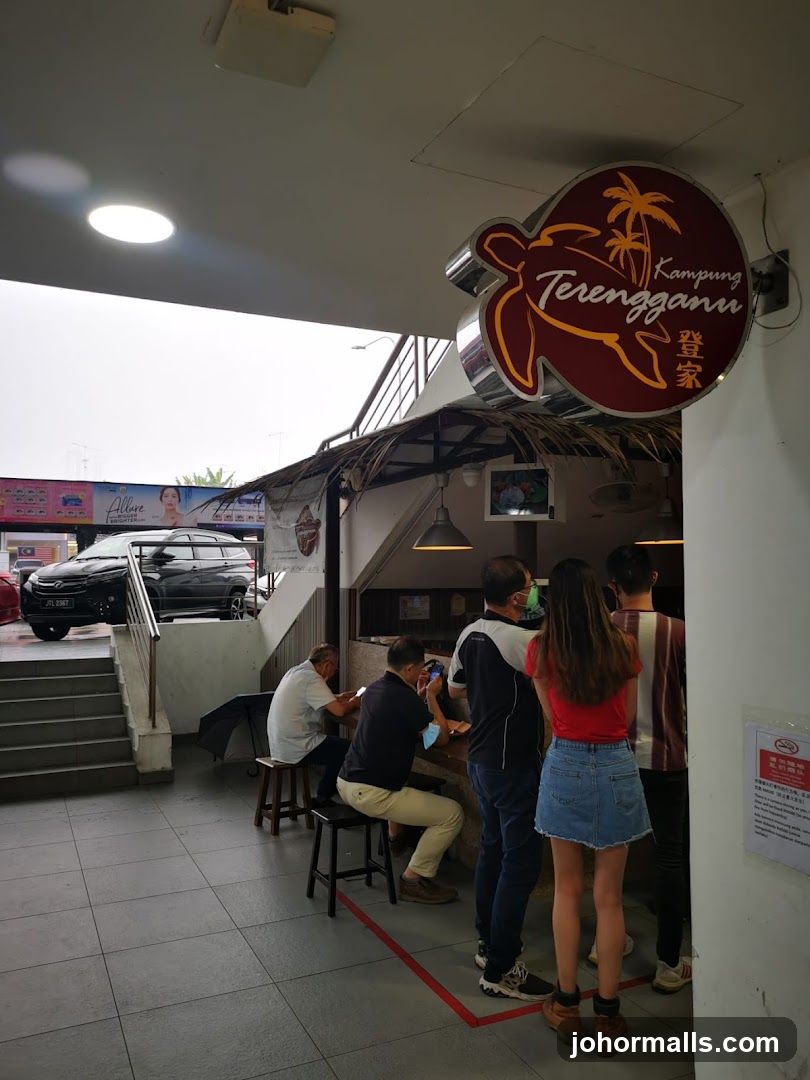 A sign for Kampung Terengganu is visible, featuring a turtle and palm trees. People are seen queuing and sitting at a counter outside the establishment. A car is parked on the left side of the image, near a set of stairs.