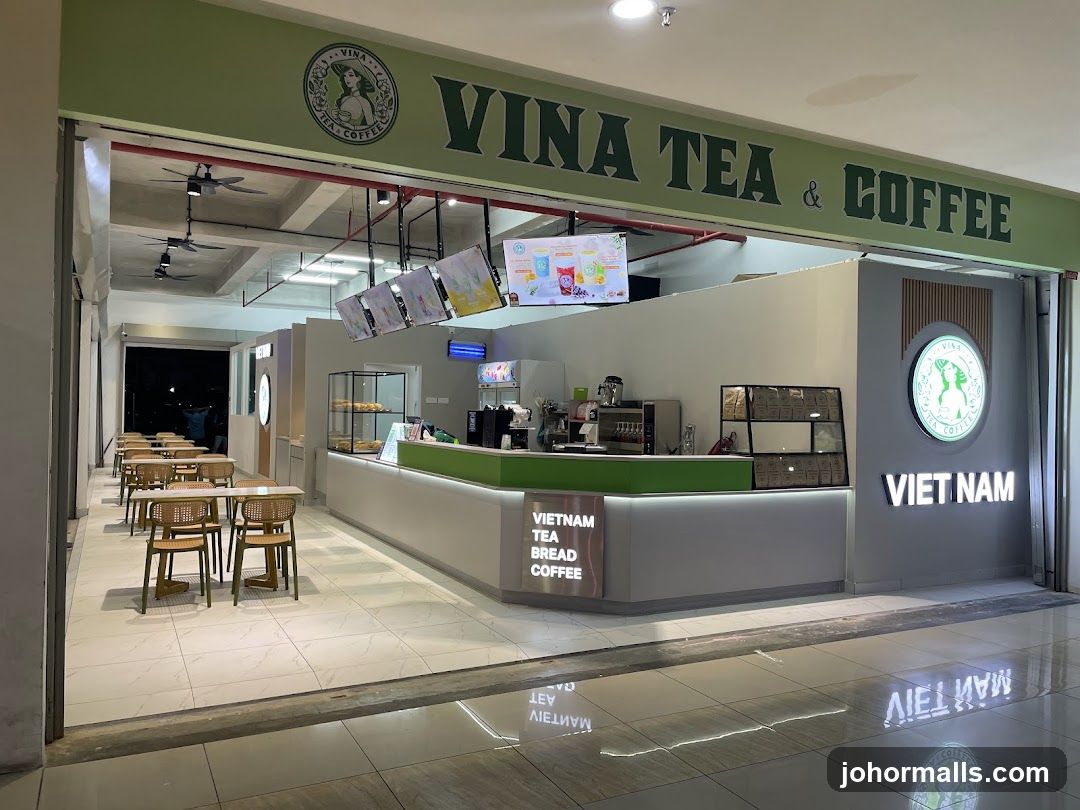 The exterior of a Vina Tea & Coffee shop. The shop has a green and white color scheme. The name "VINA TEA & COFFEE" is displayed prominently above the entrance. Inside, there is a counter with a display case, coffee machines, and seating areas with tables and chairs. The shop also has a sign that says "VIET NAM".