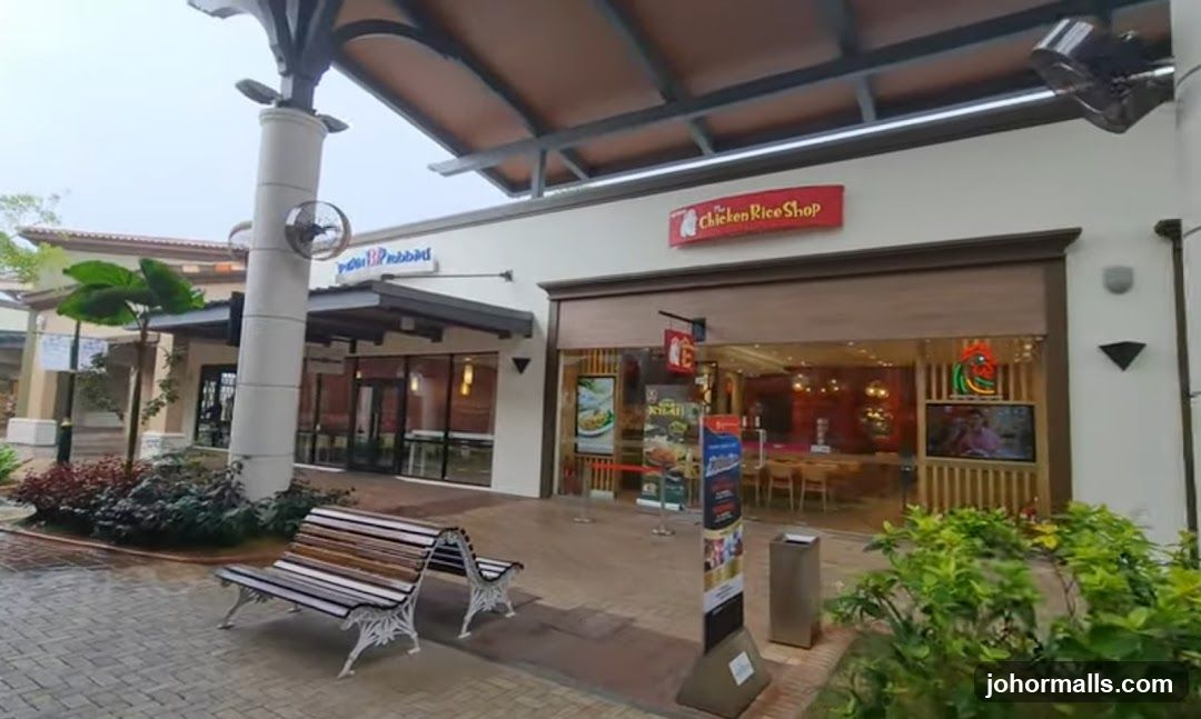 The exterior of a Chicken Rice Shop with a red sign above the entrance. To the left, another shop with a blue sign is visible. Benches and greenery are in front of the shops.