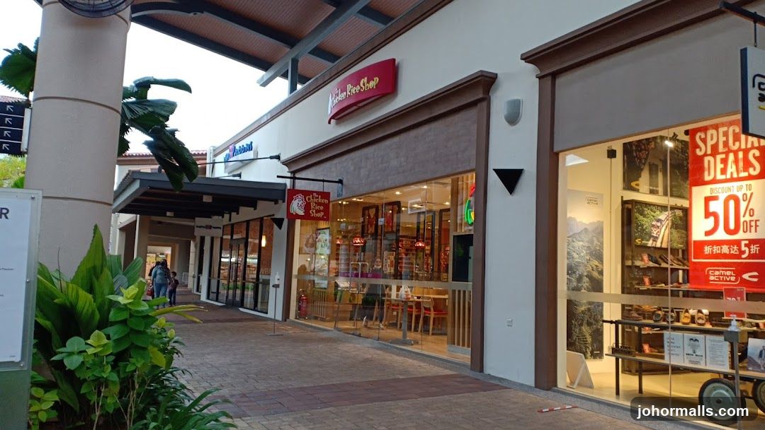 The exterior of a shopping arcade with several storefronts. One storefront is for "The Chicken Rice Shop" with its logo and name visible. Another storefront displays a "Special Deals Discount Up To 50% Off" sign for "Camel Active" shoes.