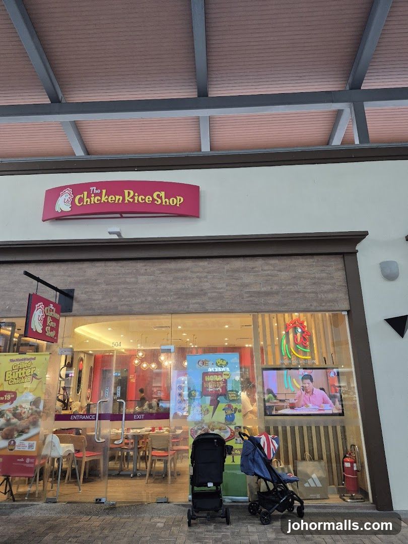 The exterior of a restaurant called "The Chicken Rice Shop". The entrance is through glass doors. Inside, tables and chairs are visible. There are also promotional posters and a television screen displaying content. Two strollers are parked outside the entrance.