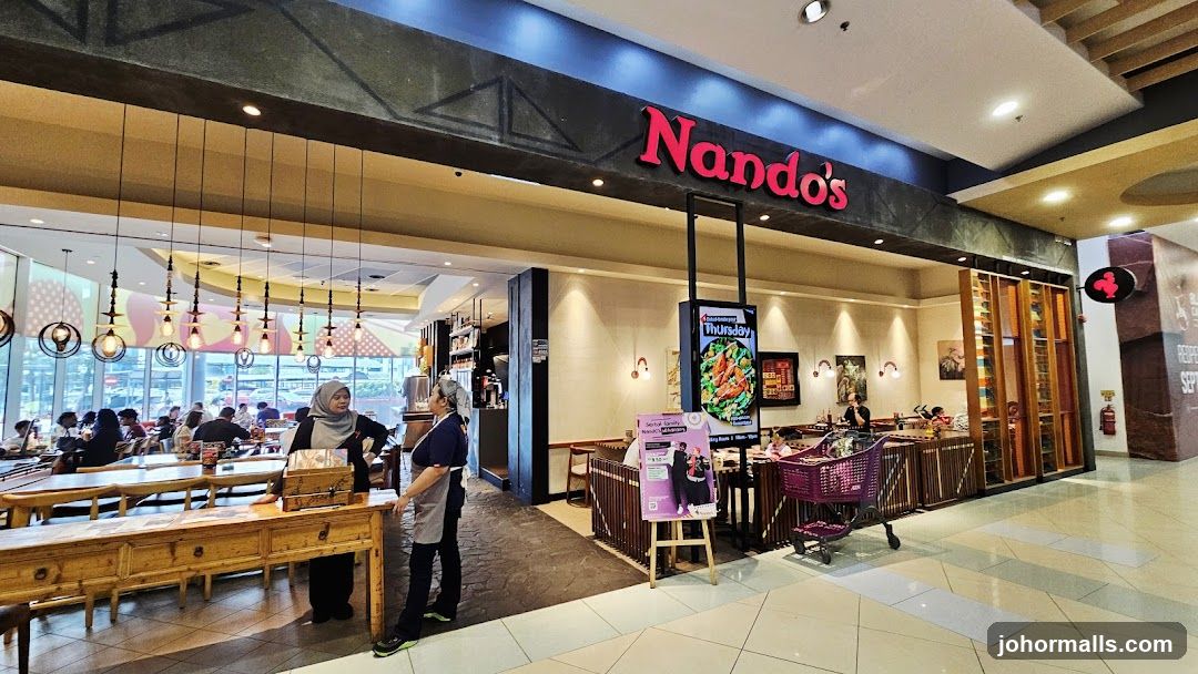 The exterior of a Nando's restaurant with its prominent red logo above the entrance. Inside, tables are set with diners seated. The restaurant features warm lighting and decorative elements. A digital display shows a food promotion, and a poster for a senior family meal deal is visible.