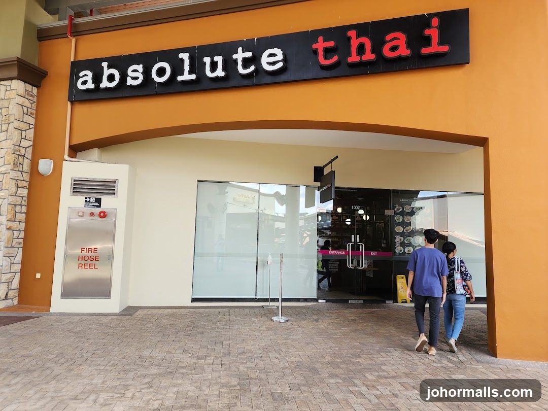 The exterior of an "Absolute Thai" restaurant. A large black sign with white and red lettering spelling out "absolute thai" is mounted above an orange archway. Below the archway is a glass storefront with double doors. The doors are marked with "ENTRANCE" and "EXIT". Two people are walking towards the entrance. To the left of the entrance is a metal cabinet labeled "FIRE HOSE REEL".