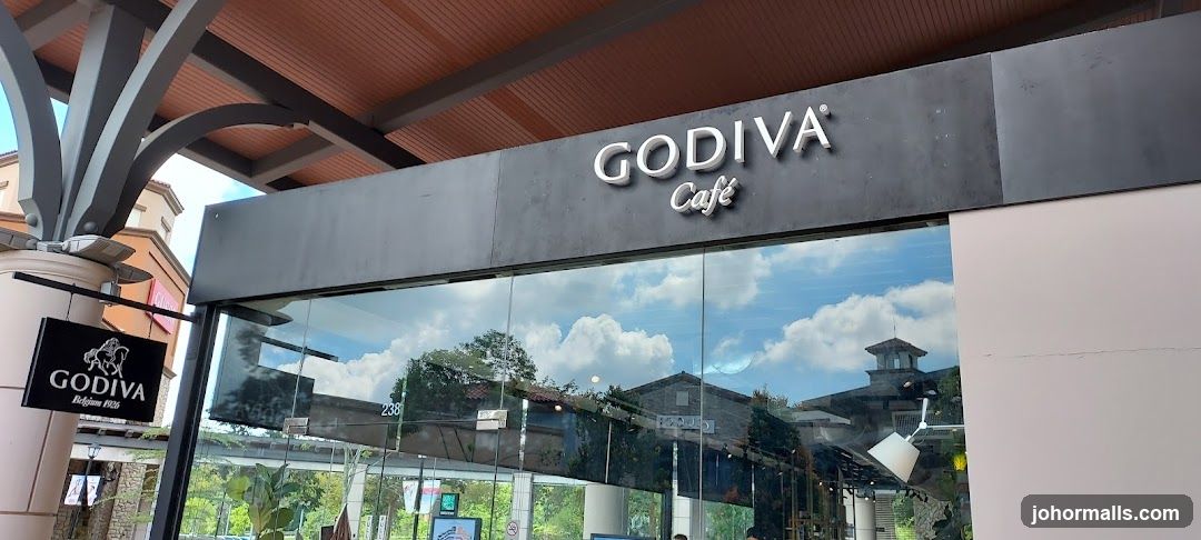 The exterior of a Godiva Cafe with a large glass window and a prominent sign above. A smaller hanging sign with the Godiva logo is visible on a pillar to the left. The reflection in the window shows buildings and trees.