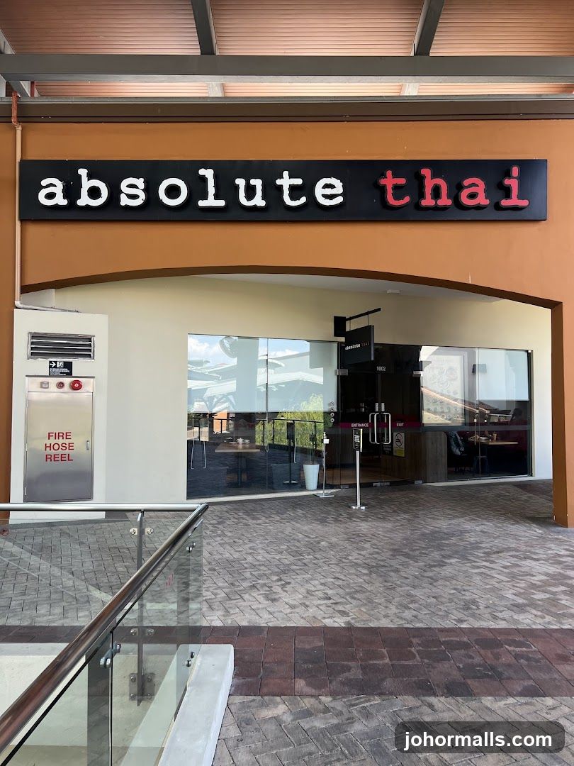 The exterior of the Absolute Thai restaurant features a prominent black sign with white and red lettering that reads "absolute thai". The entrance is through a glass door with "ENTRANCE" and "EXIT" signs. To the left of the entrance is a silver fire hose reel cabinet. The walkway in front of the restaurant is paved with brick-like tiles.