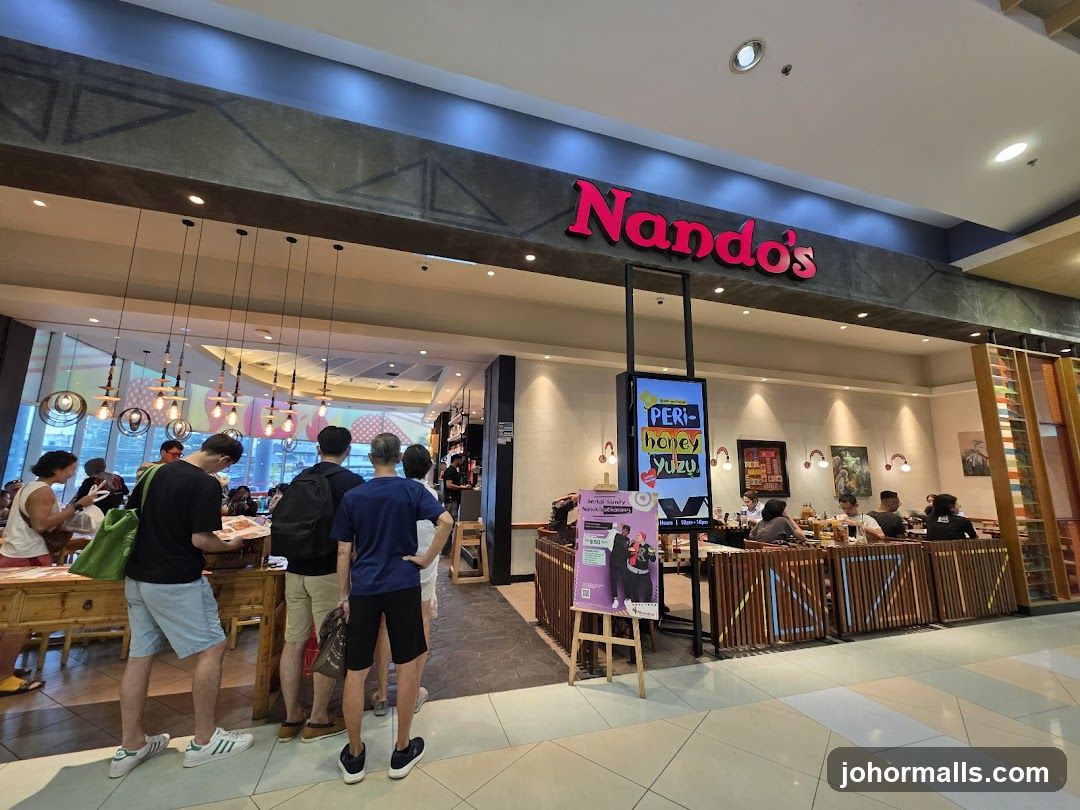 The exterior of a Nando's restaurant with its prominent red logo above the entrance. People are seen queuing and dining inside. A digital screen displays a promotion for 'Peri-honey Yuzu'.