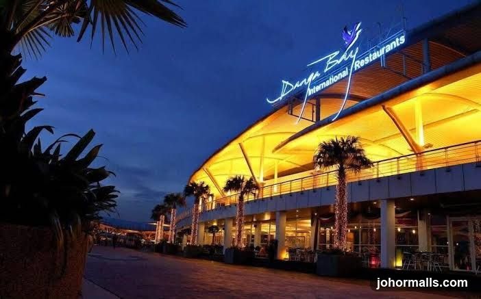 A nighttime exterior view of a modern building with a curved, illuminated roof. The building has multiple levels with balconies and large windows. Palm trees are visible in front of the building, some decorated with lights. The signage "Danga Bay International Restaurants" is prominently displayed in neon lights on the upper facade.