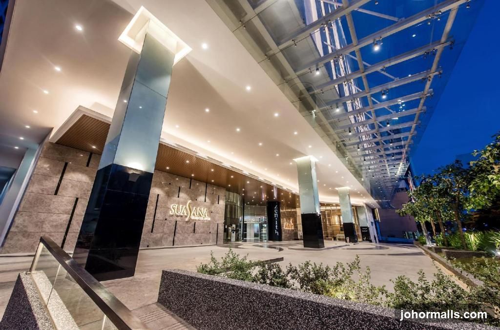 The exterior of the Suasana Hotel entrance at dusk. The building features a modern design with large pillars, a glass roof structure, and stone facade. The hotel's name is prominently displayed on the building.