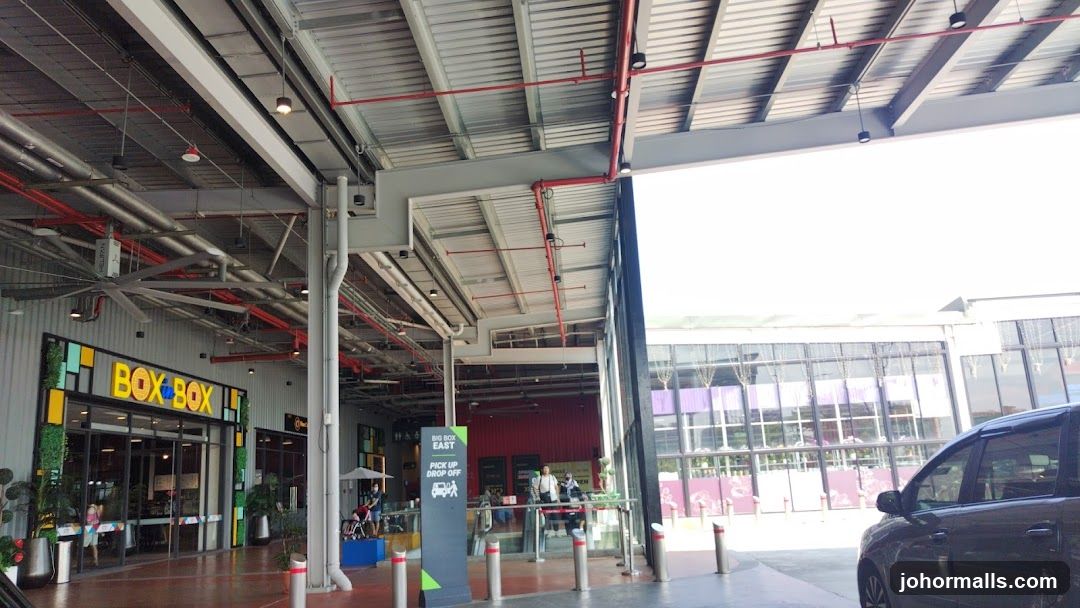 The exterior of a store with a prominent "BOX BOX" sign above the entrance. The entrance features large glass doors and windows, with some greenery and planters flanking the doorway. The building has a modern industrial design with exposed beams and metal roofing. To the right, a sign indicates "BIG BOX EAST PICK UP DROP OFF". In the background, other storefronts with large glass facades are visible.