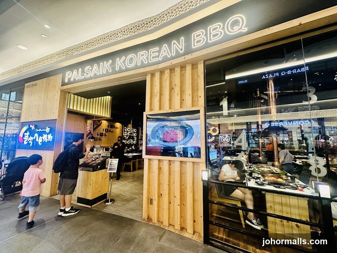 The exterior of a Korean BBQ restaurant with a prominent neon sign that reads "PALSAIK KOREAN BBQ". The entrance is framed by light-colored wood. Inside, diners are visible at tables with grills. A screen displays raw meat.