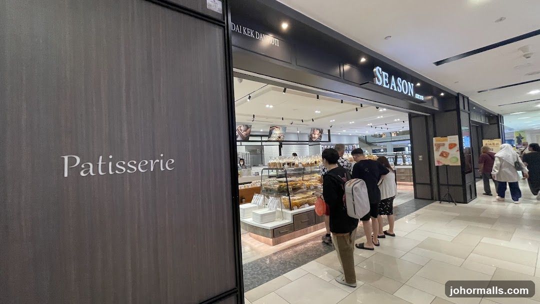 The exterior of a patisserie with a prominent "SEASON" sign above the entrance. The shop displays a variety of baked goods in glass cases. Several customers are seen queuing to enter. The word "Patisserie" is also visible on a dark panel to the left of the entrance.