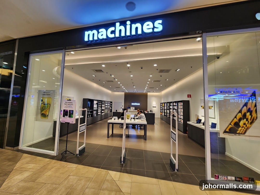 The exterior of a Machines store, an Apple Premium Reseller. The store has a wide glass entrance with the "machines" logo illuminated above. Inside, rows of Apple products are displayed on shelves and tables. Two customers are seated at a table, possibly discussing purchases with a staff member. Promotional posters for iPhones and MacBooks are visible in the windows.