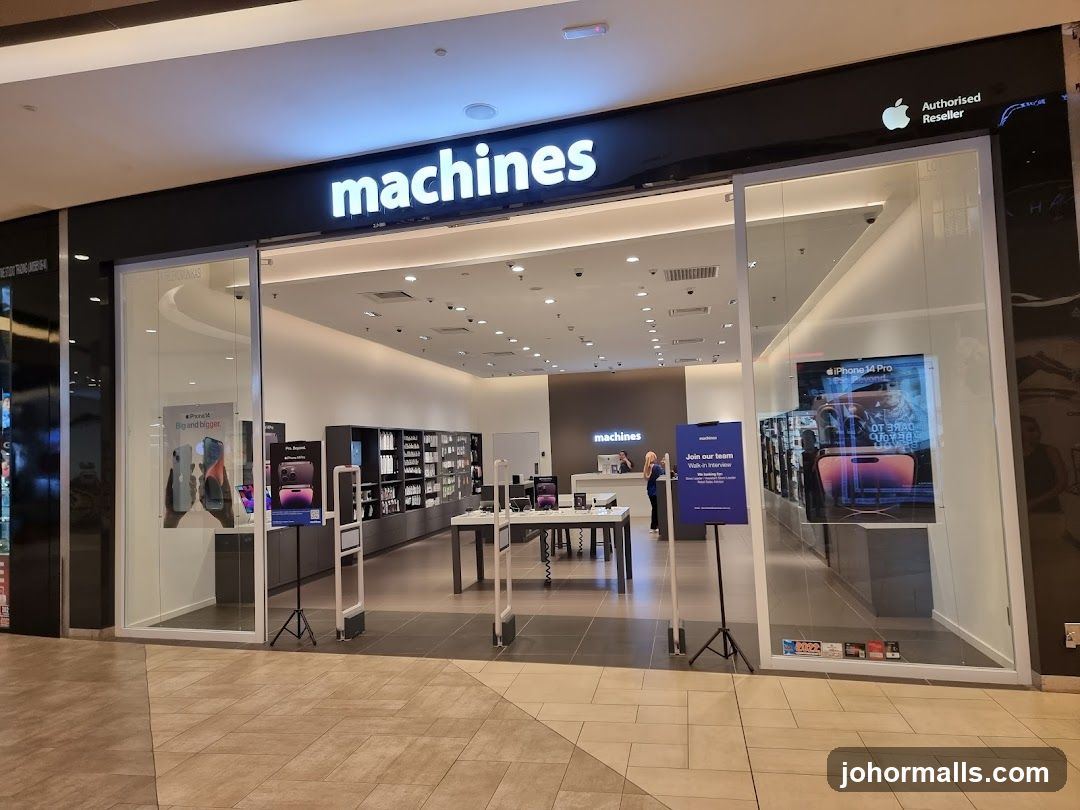 The exterior of a Machines store, an authorized Apple reseller. The store has a large glass facade with the "machines" logo prominently displayed above the entrance. Inside, Apple products are displayed on tables and shelves. An advertisement for the iPhone 14 Pro is visible on the right side of the entrance.