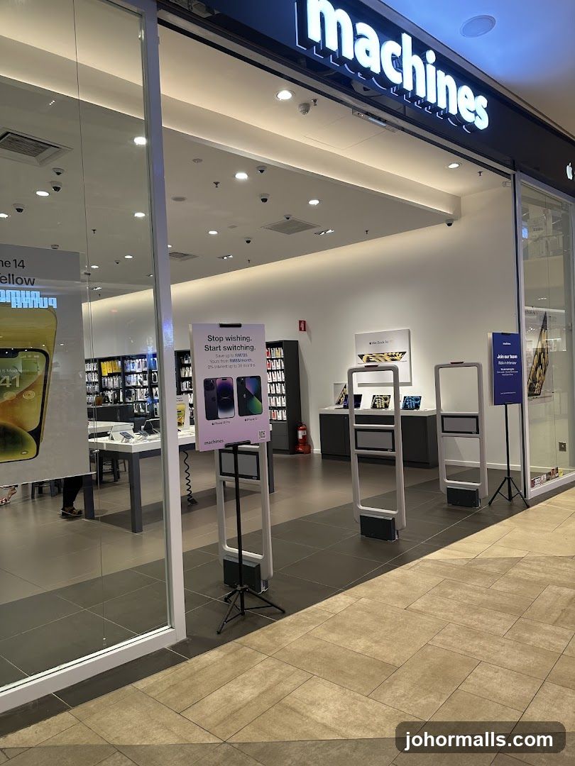 The exterior of a Machines store, an Apple Premium Reseller. The store has a large glass facade with the "machines" logo illuminated in white above the entrance. Inside, Apple products are displayed on shelves and tables. A promotional sign is visible near the entrance, advertising a "Stop wishing. Start switching." campaign with savings up to RM700. Security gates are positioned at the entrance.