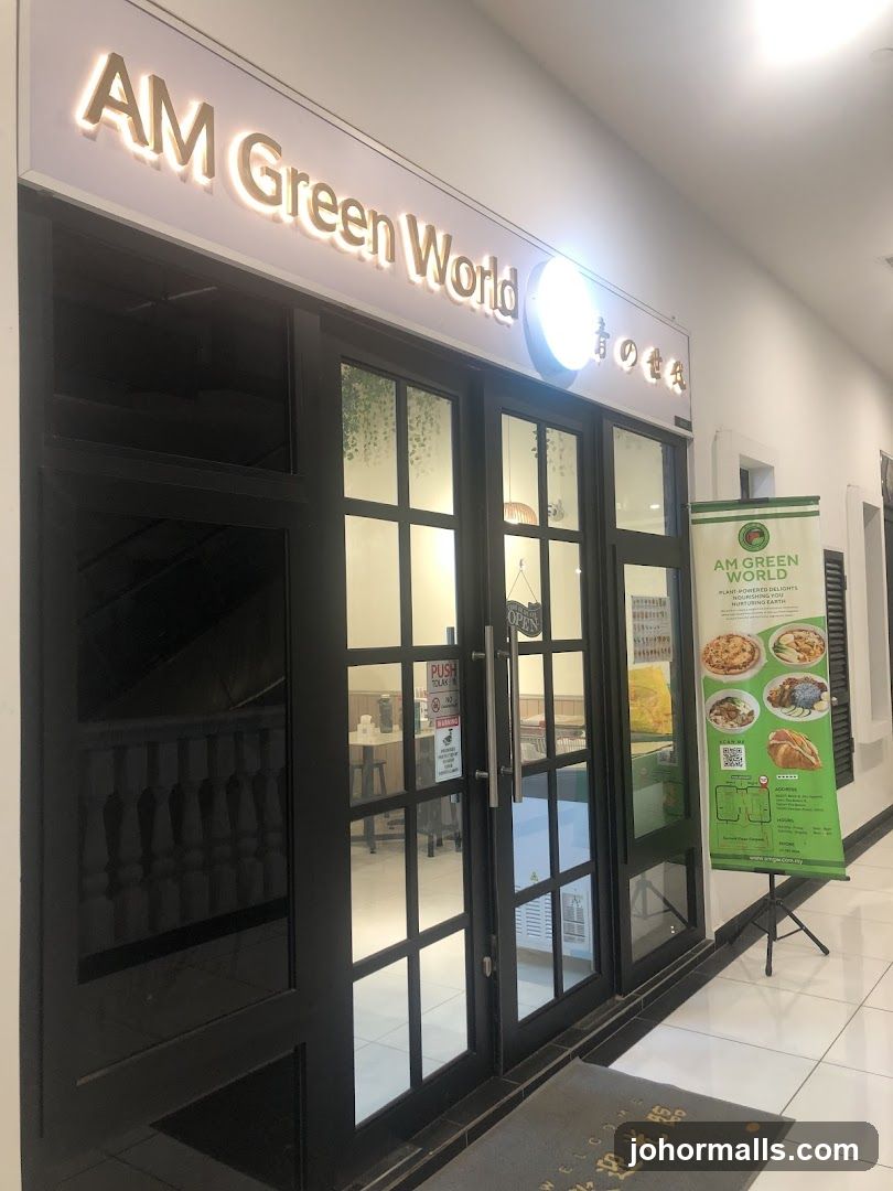 The exterior of a restaurant called AM Green World. The entrance has double glass doors with black frames. Above the doors, the name "AM Green World" is illuminated in gold lettering. To the right of the entrance, a green banner displays images of food and the text "AM GREEN WORLD PLANT-POWERED DELIGHTS NOURISHING EARTH".