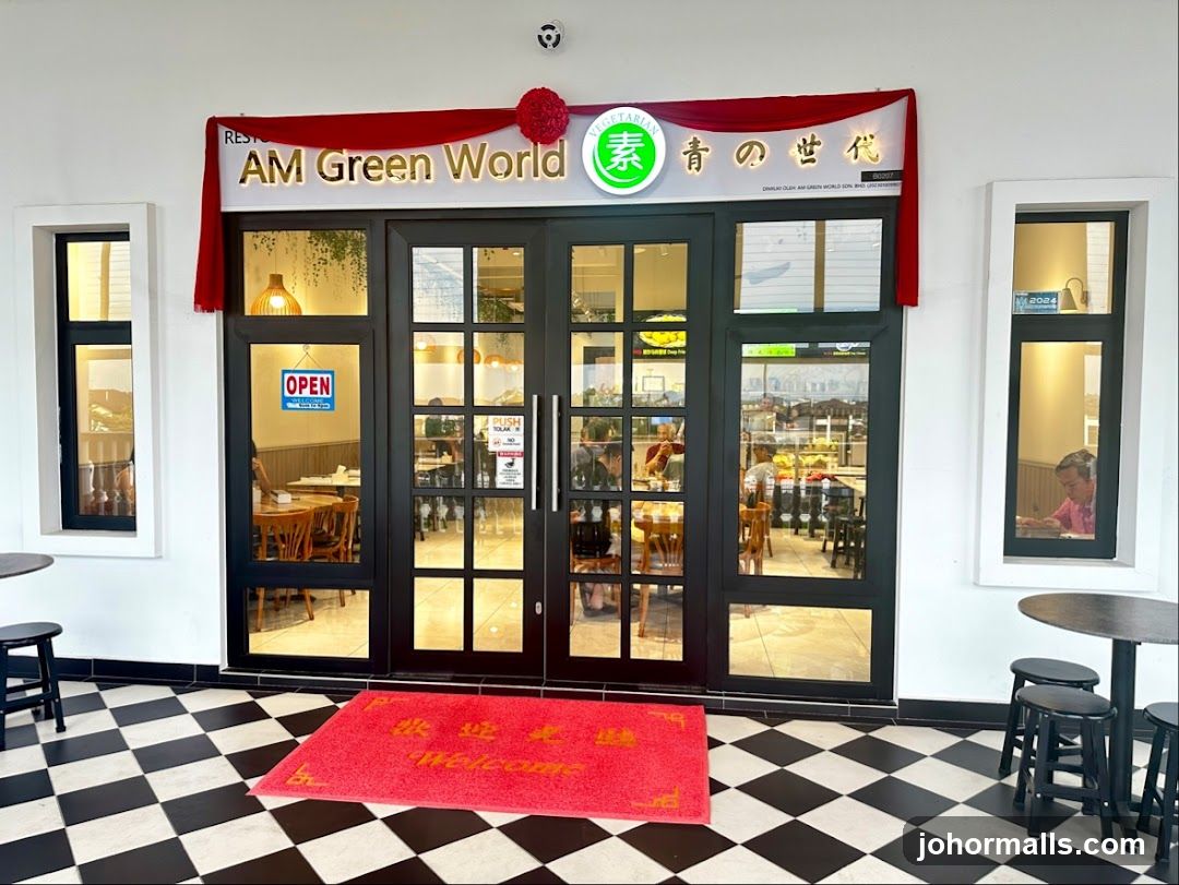 The exterior of a vegetarian restaurant called AM Green World. The entrance features double glass doors with a red welcome mat in front. There are windows on either side of the entrance, and the restaurant's name is displayed prominently above the doors. Red drapes hang on either side of the signage.