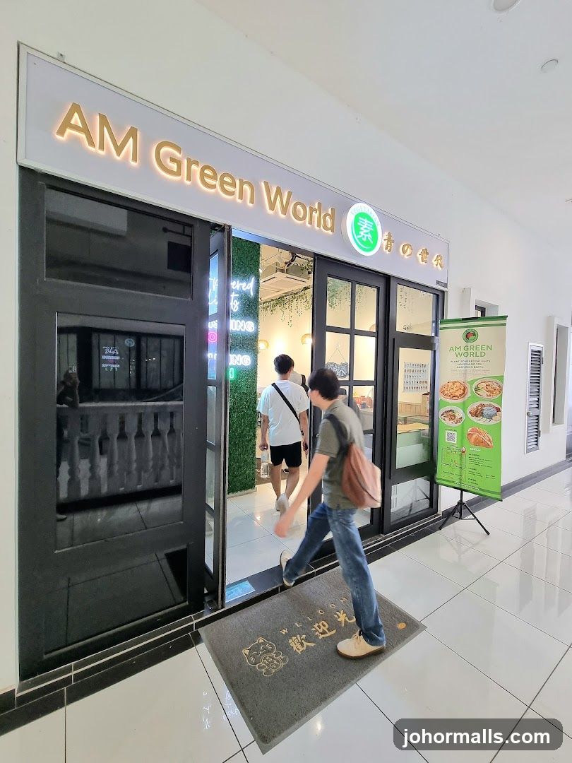 The exterior of a shop called AM Green World. The shop has a large sign above the entrance that reads "AM Green World" in gold lettering. There are glass doors and windows, and a welcome mat at the entrance. A green banner with food images and text is displayed outside the shop.