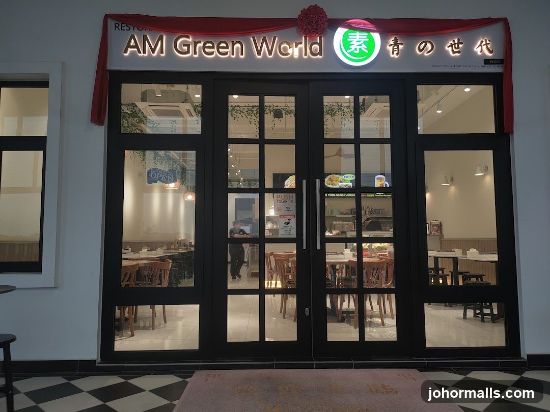 The exterior of a restaurant called AM Green World. The entrance has large glass doors with a black frame. Above the doors, the restaurant's name is displayed in illuminated letters. There are tables and chairs visible inside the restaurant through the glass. A red fabric decoration is draped above the signage.