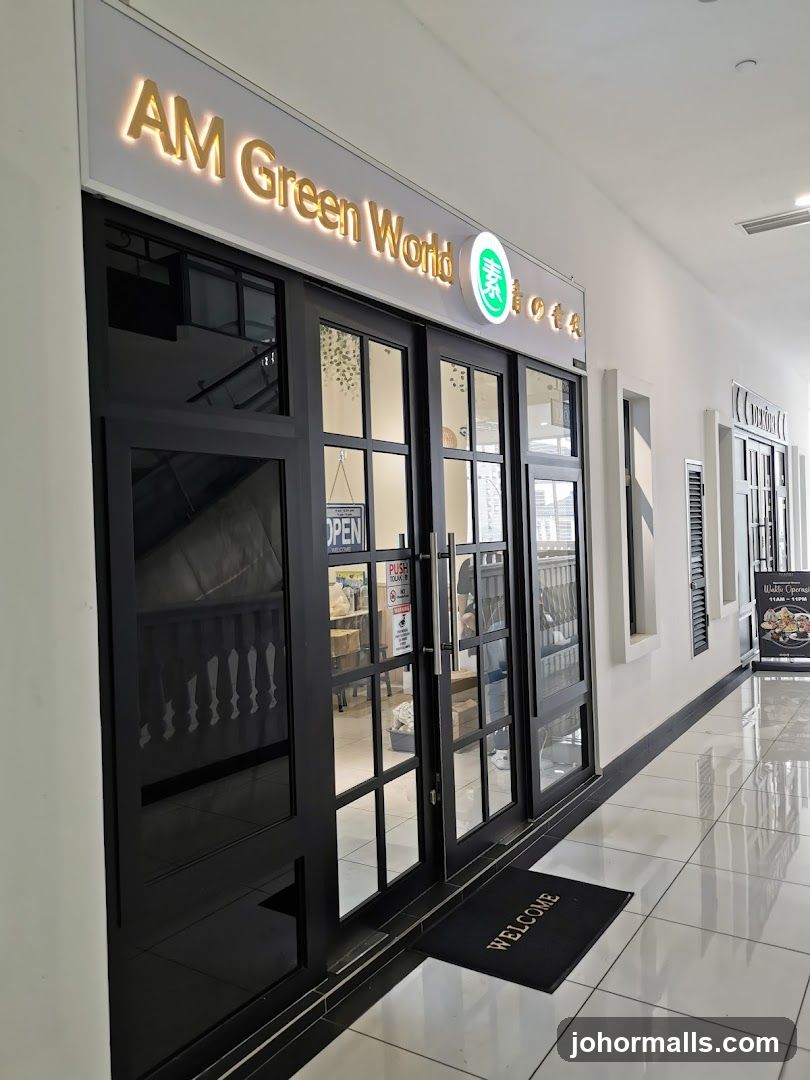 The storefront of AM Green World features a large sign with illuminated golden text and a green circular logo. The entrance consists of double glass doors with black frames, allowing a view into the interior. A black welcome mat is placed in front of the doors. To the right of the entrance, a poster displays food items and operating hours.