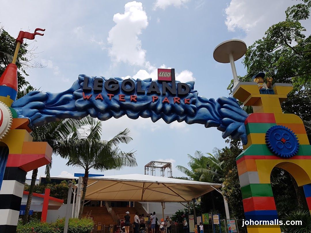 Colorful LEGO-themed archway with "LEGOLAND WATER PARK" signage, constructed from oversized LEGO bricks, flanked by LEGO structures including a gear and flagpole. Background shows palm trees, visitors near an entrance canopy, and partly cloudy sky.
