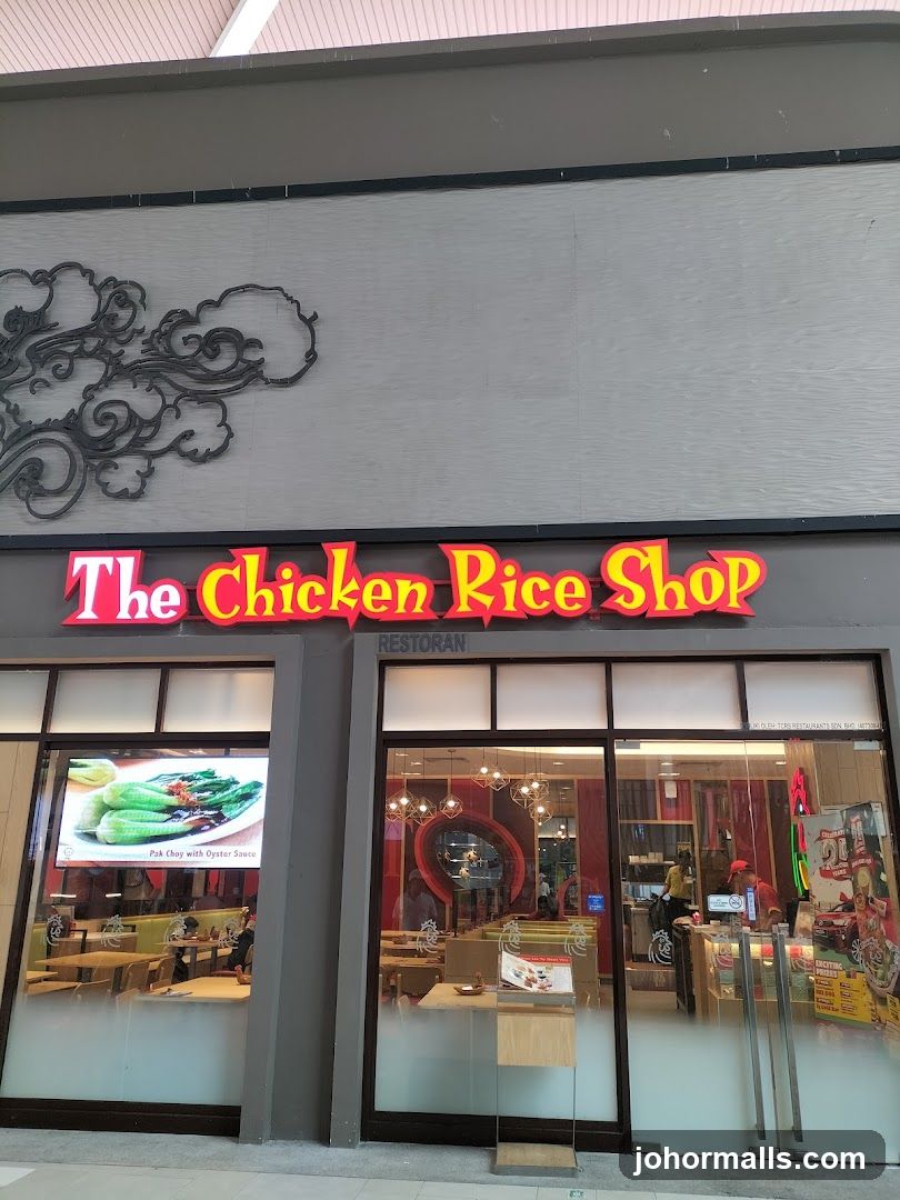 The exterior of a restaurant called "The Chicken Rice Shop". The sign is illuminated in red and yellow. Below the sign, the word "RESTORAN" is visible. The storefront has large glass windows and doors, offering a view of the interior seating area and some menu displays. One window features a digital display of a dish called "Pak Choy with Oyster Sauce".