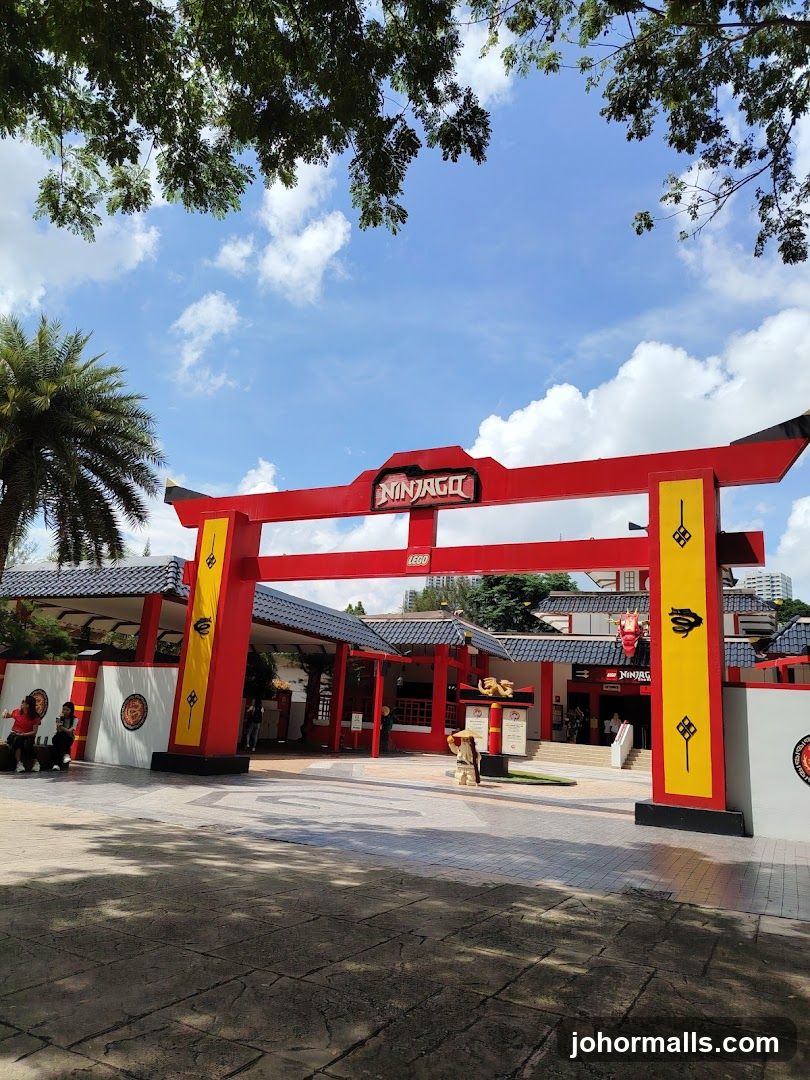 Red and yellow archway with "NINJAGO" and "LEGO" logos above entrance; traditional-style buildings behind with tiled roofs; people walking or sitting nearby; trees overhead under blue sky with clouds.