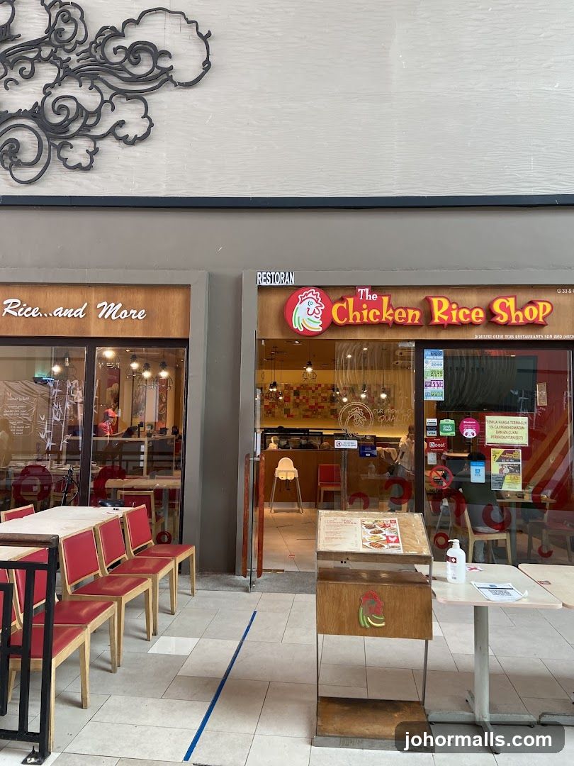The exterior of a restaurant called "The Chicken Rice Shop" is shown. The shop has a red and yellow sign with a chicken logo. Next to it is another shop with the sign "Rice...and More". There are tables and chairs outside both shops. A menu stand is placed in front of "The Chicken Rice Shop".
