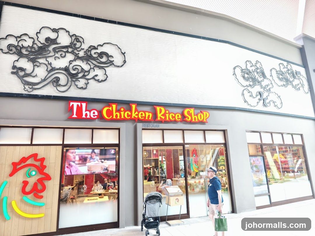 The exterior of a restaurant called "The Chicken Rice Shop". The sign is red and yellow. There is a large stylized chicken graphic on the left side of the entrance. The windows show the interior of the restaurant. There are decorative cloud motifs on the wall above the sign.