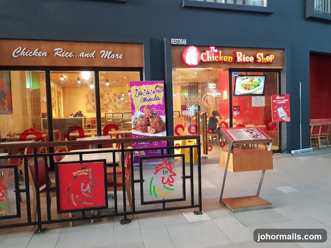 The exterior of two adjacent restaurants. The restaurant on the left is named "Chicken Rice...and More". The restaurant on the right is named "The Chicken Rice Shop". Both restaurants have glass fronts and visible interiors. There is a promotional poster for "Zazara Masala" outside the left restaurant. A menu stand is placed in front of the right restaurant.