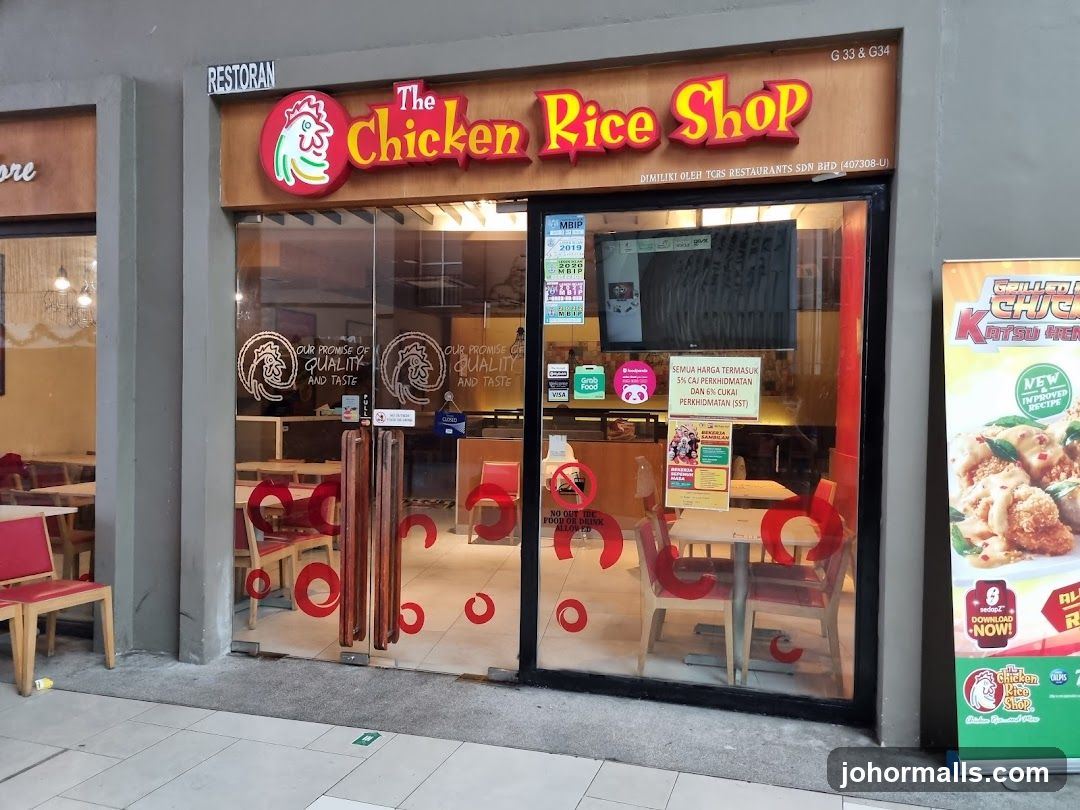 The exterior of a restaurant called The Chicken Rice Shop. The entrance has large glass doors and windows. Inside, tables and chairs are visible. A sign above the entrance reads "RESTORAN The Chicken Rice Shop". There are also various stickers and posters on the glass doors and windows. A promotional banner for "Grilled Chicken Katsu" is displayed to the right of the entrance.