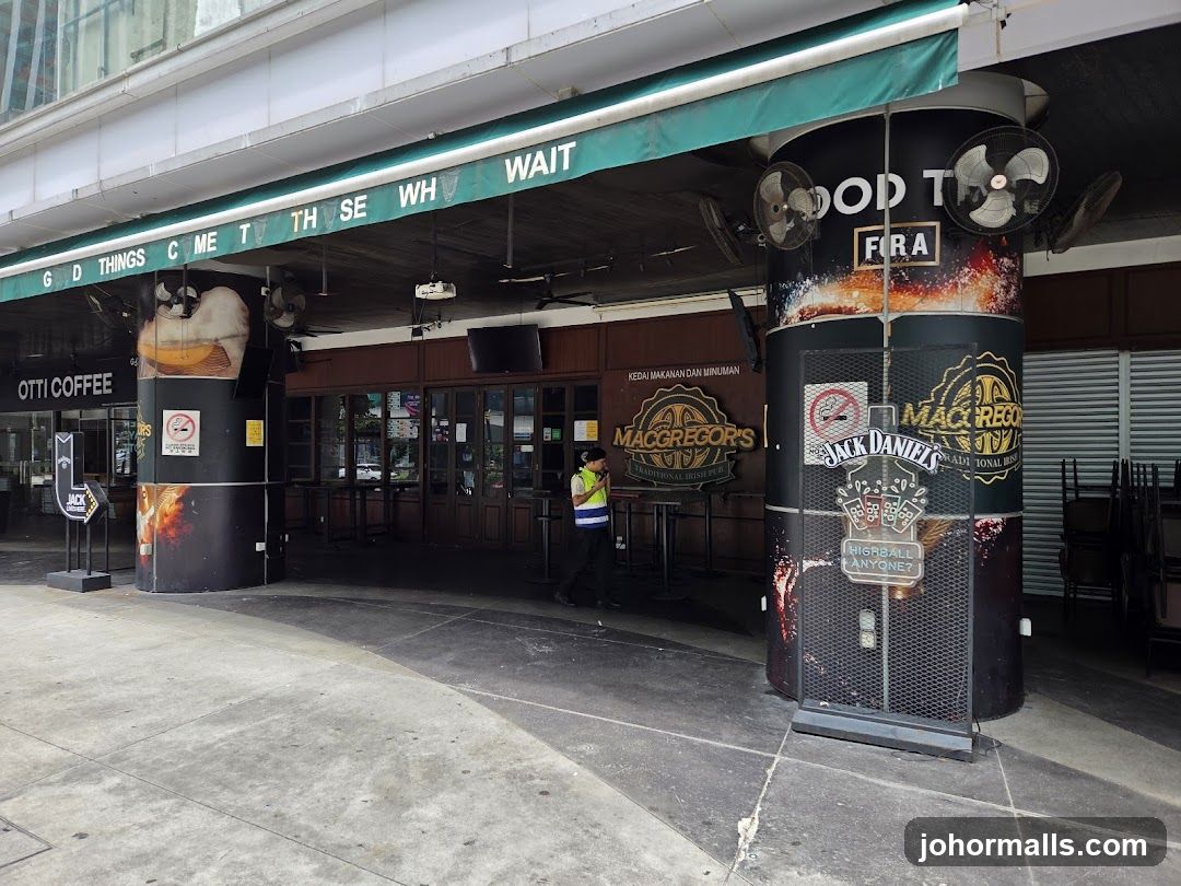 Exterior view of a pub with a green awning displaying reversed text 'GOD THINGS COME TO THOSE WHO WAIT'. A staff member in a high-visibility vest walks near the entrance. Two large cylindrical pillars are wrapped in branded advertisements featuring Jack Daniel's and MacGregor's logos. The building facade includes glass doors, signage reading 'KEDAI MAKANAN DAN MINUMAN', and an Otti Coffee sign on the left pillar.