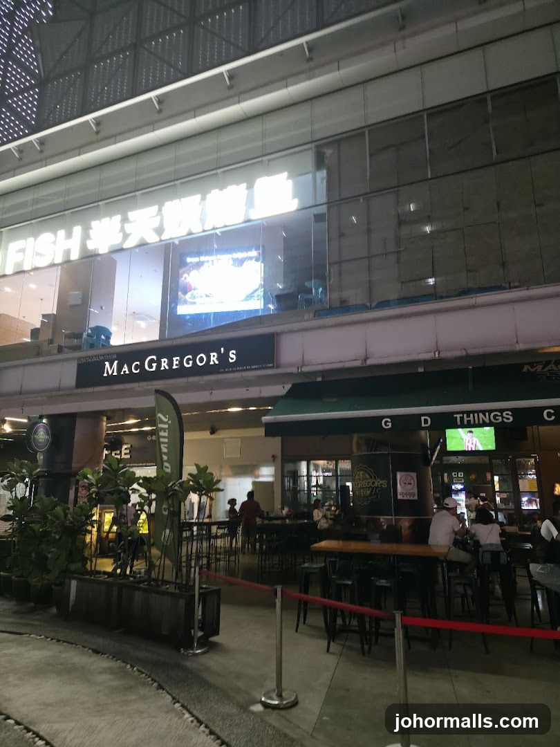 Exterior view of a multi-level commercial building with illuminated signage reading 'FISH 半天飲檳榔' above the entrance to MacGregor's. The facade includes glass windows and metal framing. Below, outdoor seating areas are visible under green awnings with 'G D THINGS C' signage. Red stanchions with rope demarcate an area near potted plants.