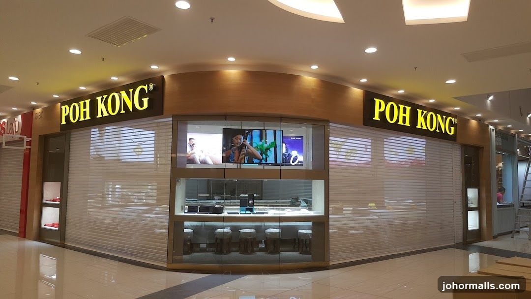 A corner storefront with illuminated "POH KONG" signage on both sides of the curved facade. The shop has glass display windows with horizontal blinds partially covering them. A central display case is visible through the glass, showing products under lighting. Above the display, a digital screen shows promotional images. The floor is tiled and reflective. No doors are open; the store appears closed or not in operation.