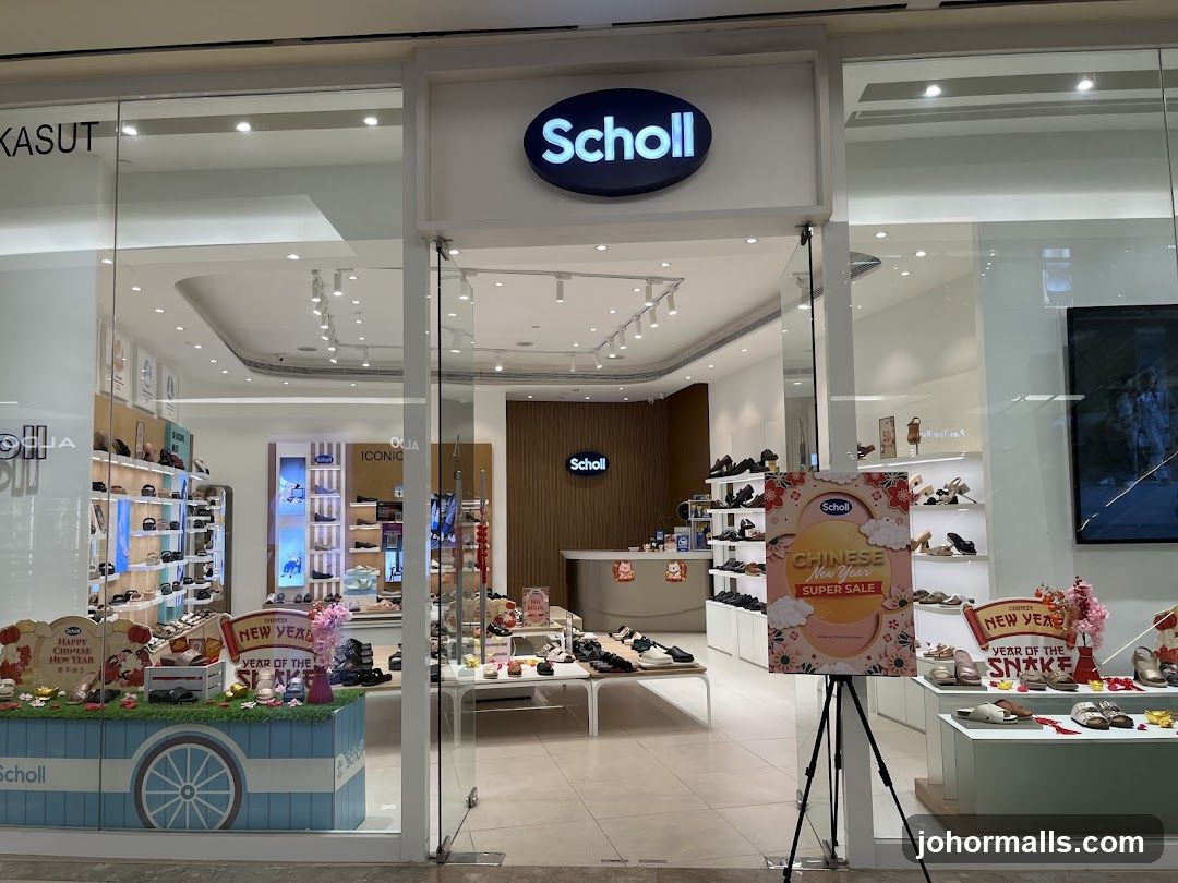 Storefront with glass doors and windows displaying interior. Above the entrance is a glowing blue oval sign with 'Scholl' in white letters. Inside, shelves of shoes are visible, along with a counter and wooden accent wall also bearing the Scholl logo.