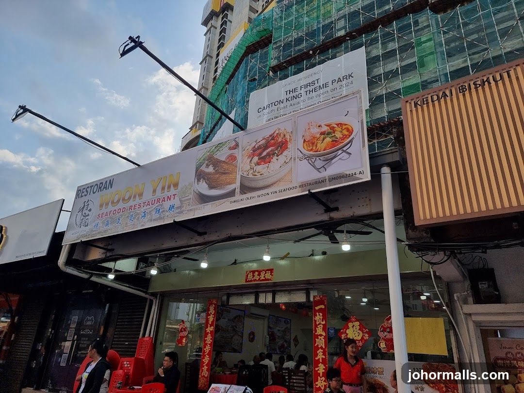 Exterior storefront with glass windows and red seating. Signage above reads 'RESTORAN WOON YIN SEAFOOD RESTAURANT'. Red Chinese New Year decorations adorn the entrance. Customers are seated inside, staff is visible. Above the shop, a large promotional banner advertises 'THE FIRST CARTON KING THEME PARK' and images of seafood dishes.