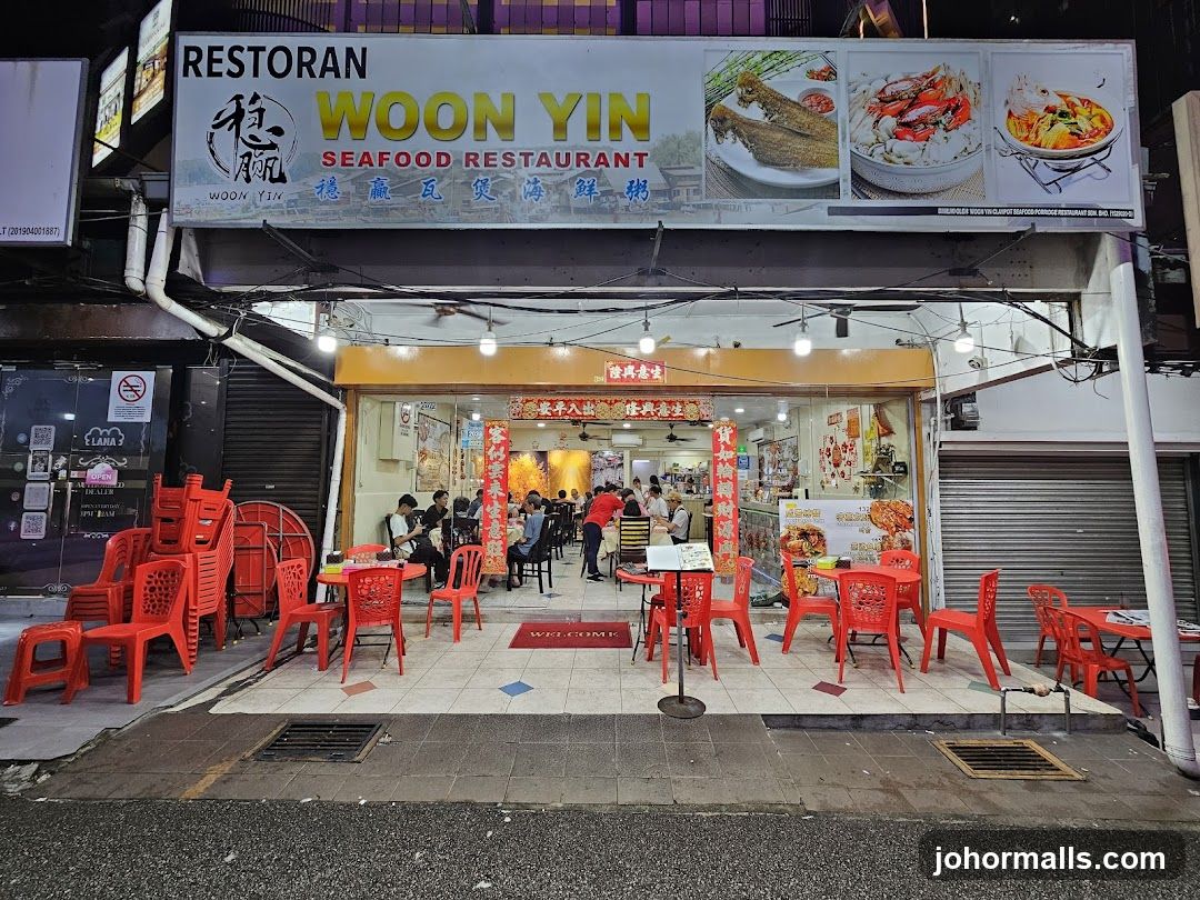 Exterior storefront with large signboard displaying "RESTORAN WOON YIN SEAFOOD RESTAURANT" in English and Chinese characters. Red plastic chairs and tables are arranged outside. Glass doors show interior dining area with patrons, red decorative banners, and a "WELCOME" mat on the floor.