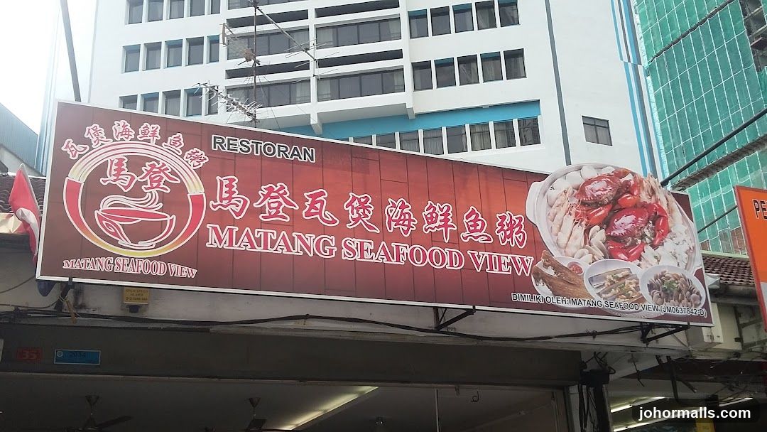 Exterior storefront with large red signboard displaying "RESTORAN MATANG SEAFOOD VIEW" in English and Chinese characters. Sign includes logo of a bowl with seafood and an image of a seafood dish on the right side. Building facade behind is modern, multi-story structure with white walls and blue accents. A Malaysian flag is partially visible to the left. No menu or interior view shown.