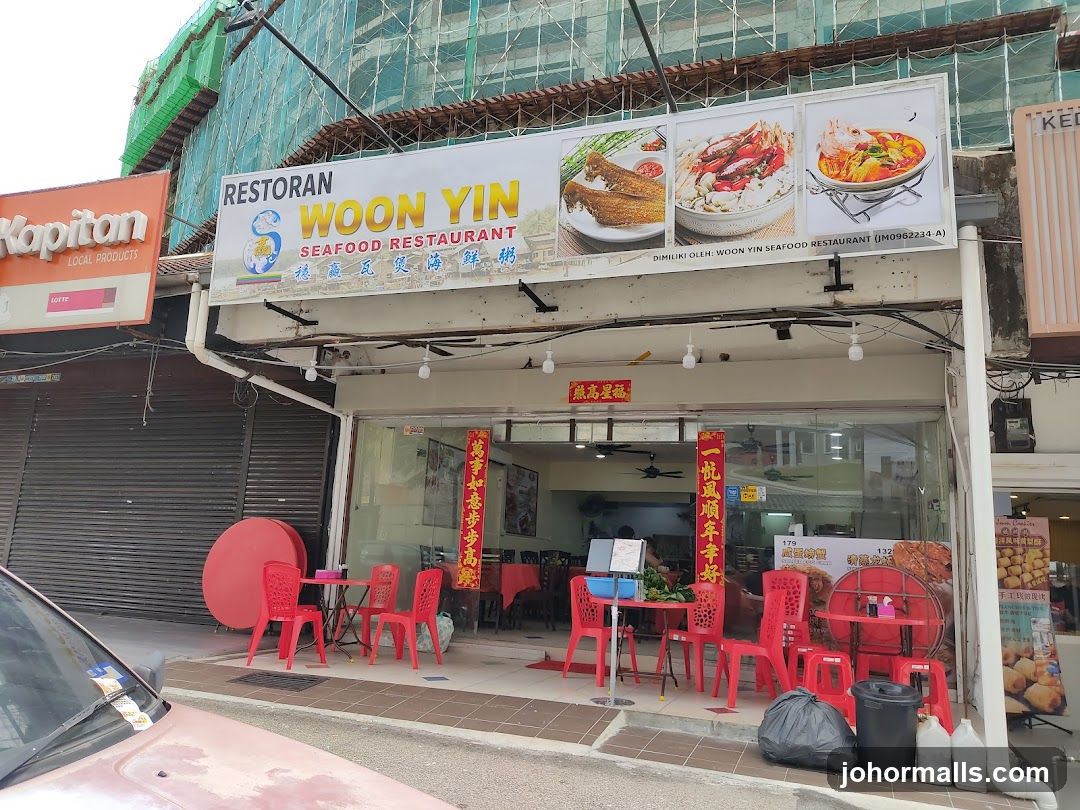 Exterior storefront with large signboard displaying "RESTORAN WOON YIN SEAFOOD RESTAURANT" in English and Chinese characters. Red plastic chairs and tables are set outside the entrance. Glass doors and windows show interior seating. Above, a green-scaffolded building is under construction. A red circular object sits on pavement near entrance. Sign includes registration number (JM0962234.A).