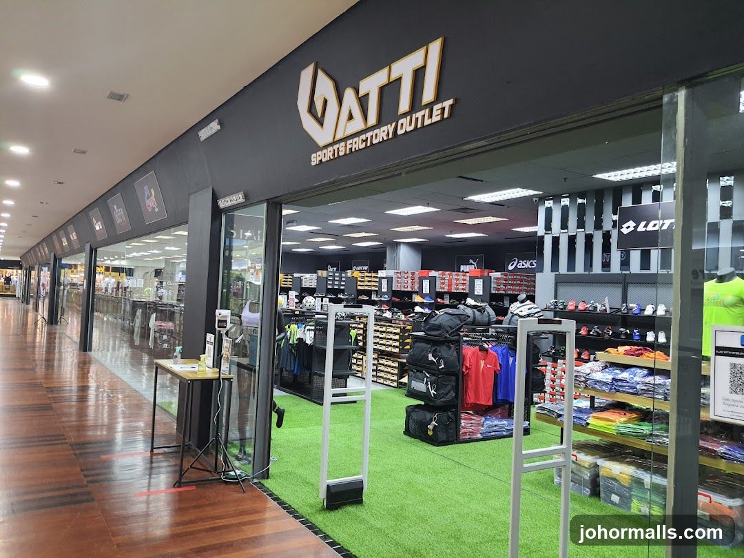 Storefront with black facade and gold lettering displaying 'VATTI SPORTS FACTORY OUTLET'. Glass doors lead into a brightly lit interior with artificial green turf flooring. Inside, racks of sports apparel and shelves of footwear are visible. A small table with a sanitizer station stands outside the entrance.