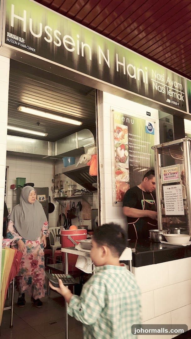 Photo of a food stall entrance with signage above the counter reading 'Hussein N Hani' and listing 'Nasi Ayam', 'Nasi lemak'. Inside, a woman in hijab stands near the counter while a man works behind it. A child holds a payment device. The interior shows kitchen equipment and a vent hood.