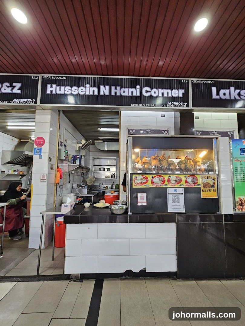 A food stall with a black sign displaying "Hussein N Hani Corner" above the counter. The interior shows a kitchen area with white tiled walls, stainless steel appliances, and a glass display case filled with roasted chickens. A staff member in a red apron is visible on the left side of the stall. The ceiling has red paneling and two bright overhead lights.