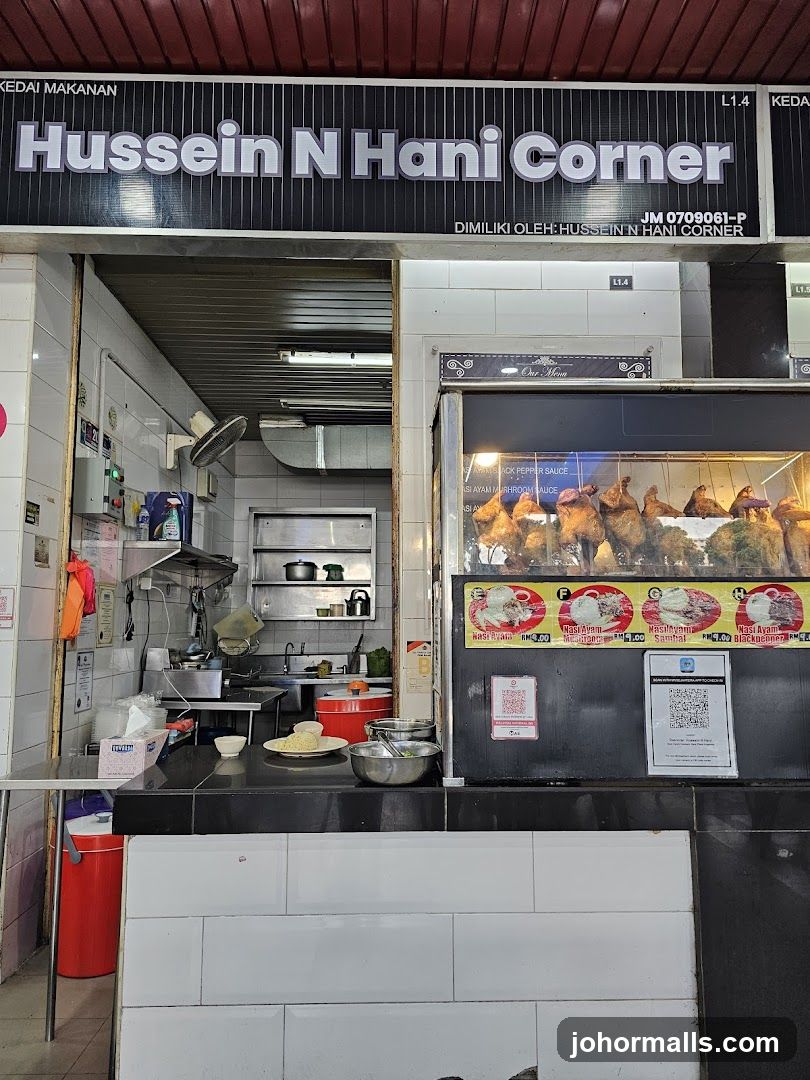 Exterior storefront of a food stall with black and white signage displaying the shop name 'Hussein N Hani Corner' above the entrance. The interior is partially visible, showing a kitchen area with stainless steel appliances, a fan, and a red trash bin. A glass display case on the right showcases roasted chickens with menu items advertised below it.