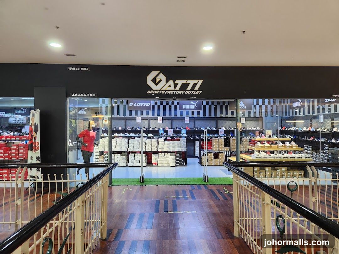 Storefront with black signage displaying 'GATTI SPORTS FACTORY OUTLET' in gold lettering above glass windows. Inside, shelves display shoe boxes and branded logos including Lotto, Puma, Adidas. A mannequin wearing a red shirt stands near the entrance. The view is from an upper level with wooden flooring and metal railings.