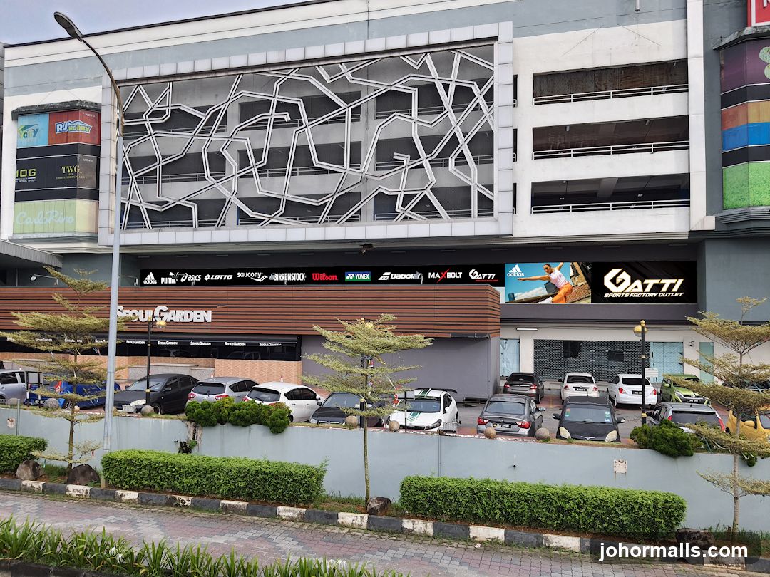 Exterior facade of a multi-level commercial building with prominent signage for 'FOUR GARDEN' and 'GATTI SPORTS FACTORY OUTLET'. Above the ground floor, a large digital display shows an athlete in motion. The upper levels feature a geometric metal screen design and multiple brand logos including Adidas, Puma, Asics, Lottu, Yonex, Babolat, Max Bolt, and Wilson. A parking lot is visible below with several cars parked near low green hedges and a paved sidewalk.