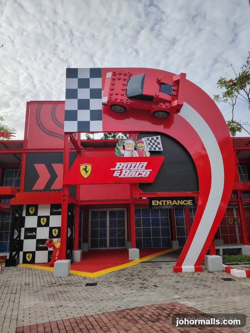 Red and white themed entrance with large curved archway featuring a Lego Ferrari car model on top, checkered flag design, and 'Build & Race' sign with Ferrari logo. Yellow 'ENTRANCE' sign below. Checkered pattern walls with Ferrari logos. Paved ground with red and grey bricks.