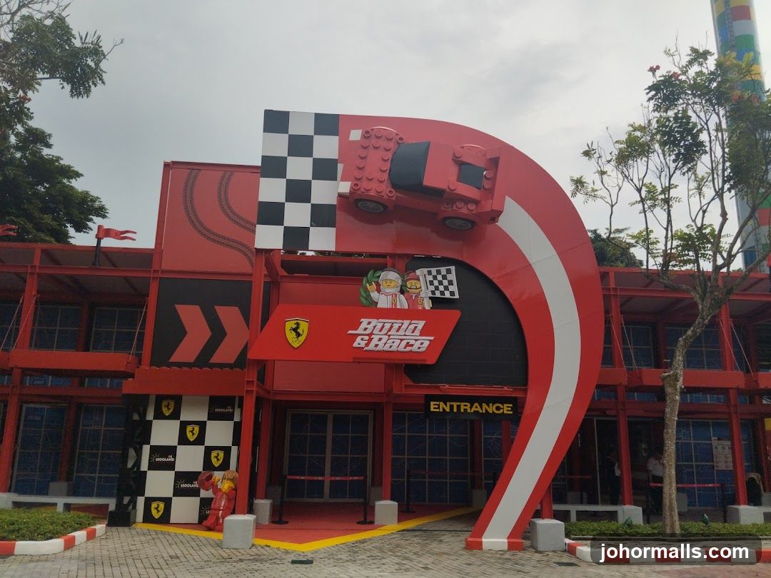 Red and black themed building facade with large curved archway featuring a Lego Ferrari car model on top. Signage reads 'Build & Race' with Ferrari logo above the entrance. Yellow 'ENTRANCE' sign below. Checkered flag pattern decorates walls and pillars. Red, white, and yellow accents throughout. Trees and paved ground in foreground.