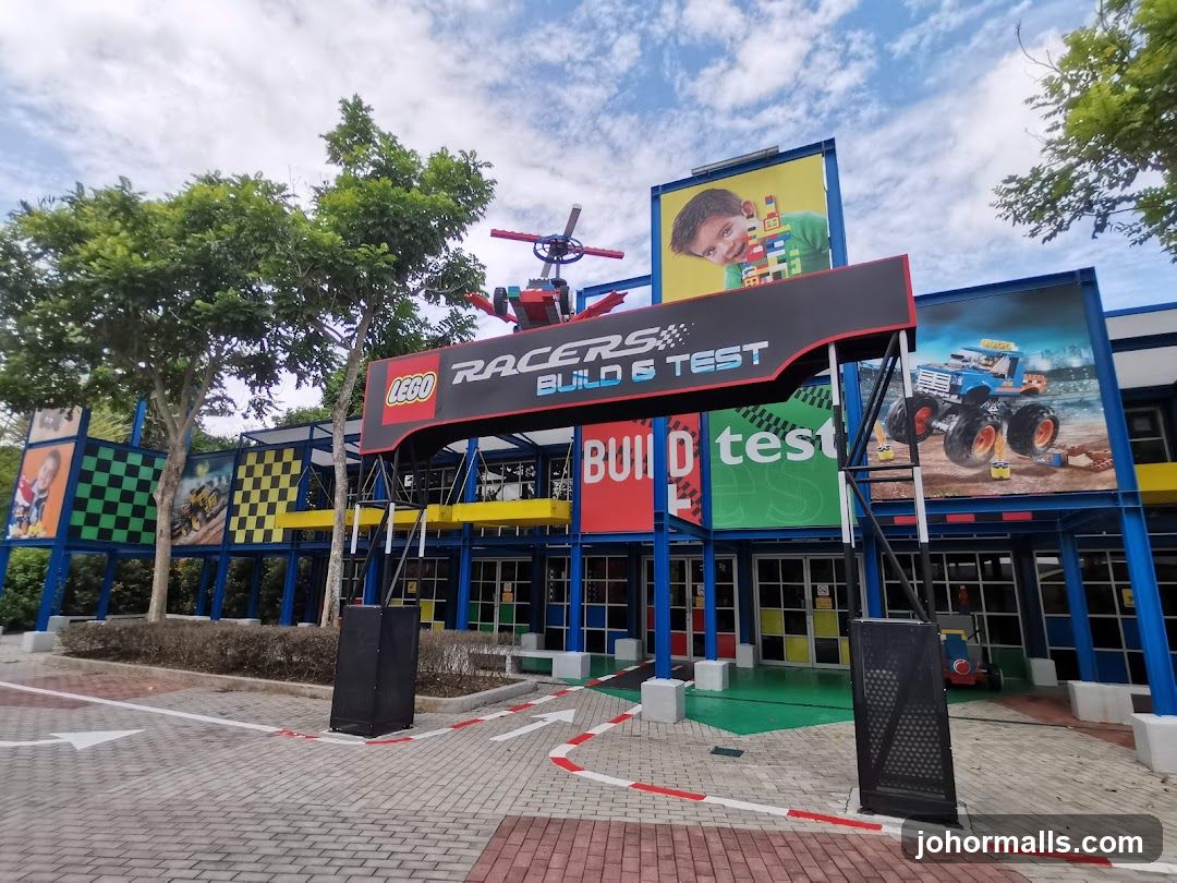 Exterior view of a colorful building facade with prominent LEGO branding and 'Racers Build & Test' signage above the entrance. The structure features blue, yellow, and red accents with large promotional graphics on walls showing children and vehicles. A checkered flag pattern decorates one side, and the ground has painted racing-style arrows and lines. Trees flank the building under a partly cloudy sky.