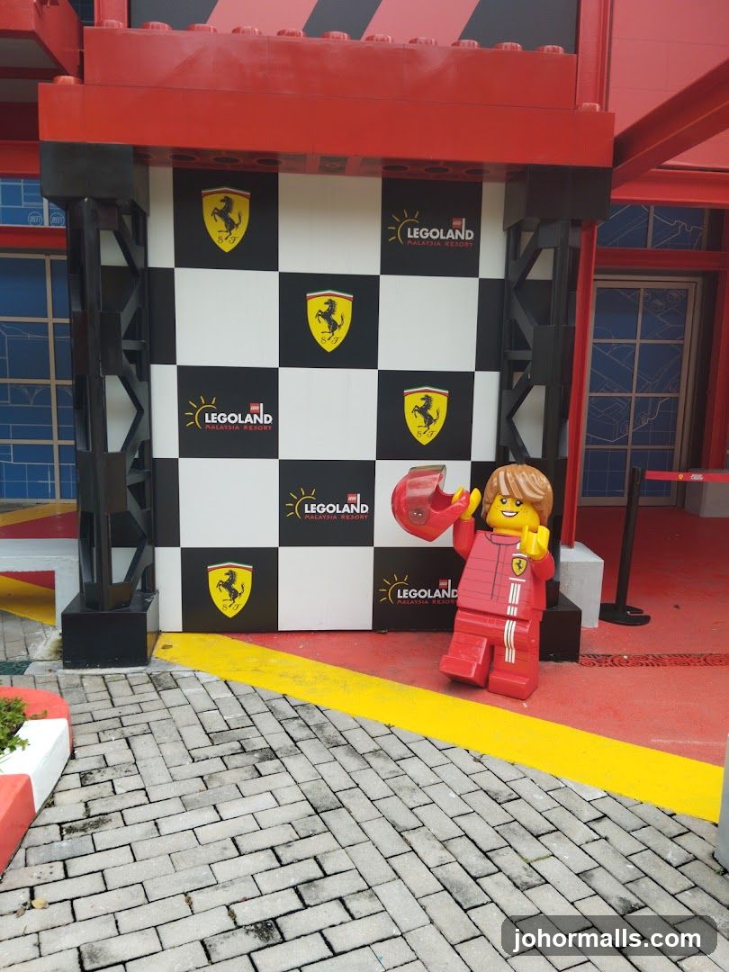 Red Lego-themed building facade with black-and-white checkered wall featuring multiple Ferrari logos and "Legoland Malaysia Resort" branding. A large red Lego figurine in a racing suit holds a helmet beside the entrance. The ground is paved with grey bricks, bordered by yellow lines. Red structural elements frame the top of the entrance.